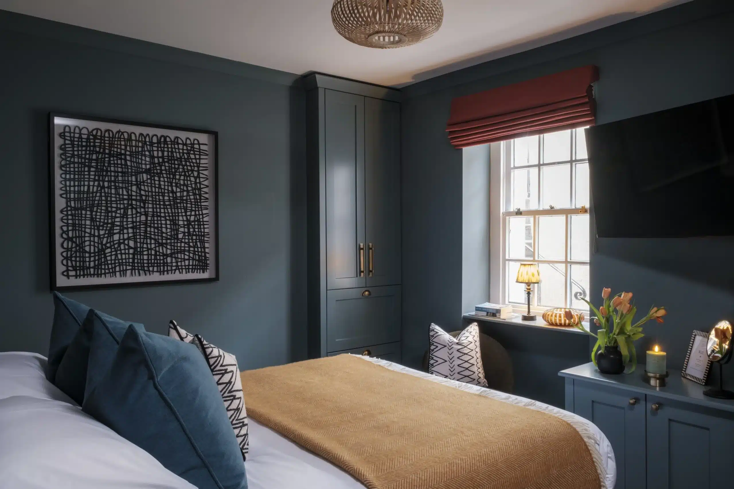 Stylish bedroom with teal walls, bed with white linens and mustard throw, black abstract wall art, and warm ambient lighting.